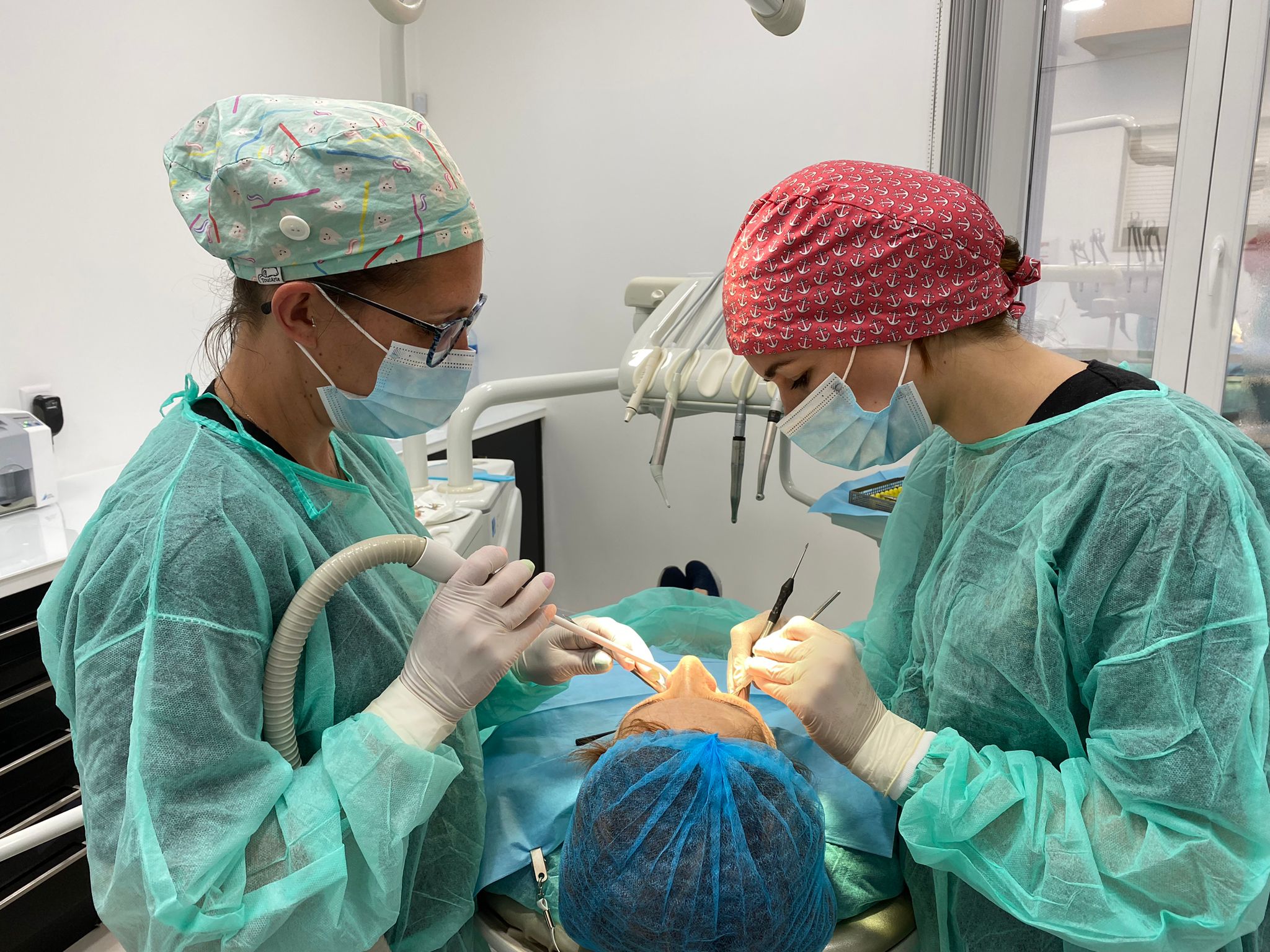 uma médica dentista e uma assistente dentária equipadas com batas cirúrgicas verdes descartáveis, toucas laranja e verde e máscaras azuis a realizar um procedimento cirúrgico numa paciente